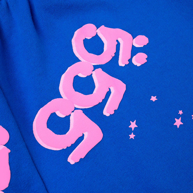 A close-up of 100% Cotton Sp5der Sweatpants shows unisex blue jogger pants with pink logo and star accents, including stylized 9:59 numbers, highlighting their bold streetwear style and relaxed fit.