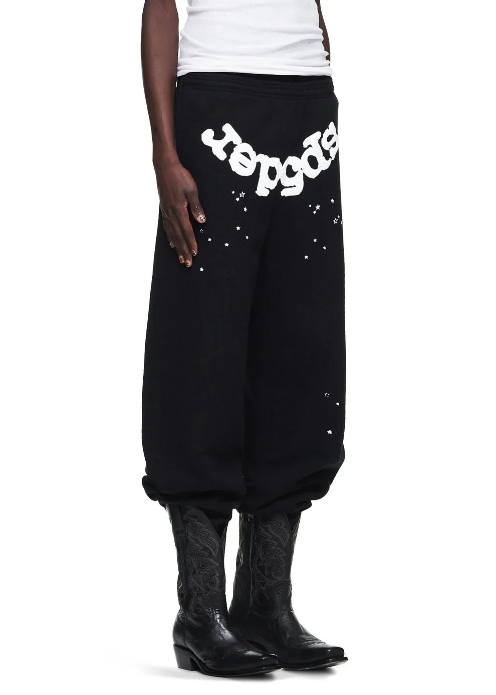 A person sports Sp5der Sweatpants—unisex black cotton pants with a front logo and star print—gathered at the ankles, styled with a white top and black cowboy boots for standout streetwear appeal.
