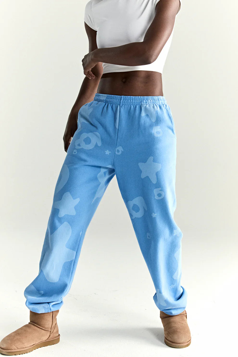 A person in a white crop top, Sp5der Light Blue All-Over Print Sweatpants 100% Cotton Unisex Streetwear Joggers, and tan UGG-style boots stands against a plain light background.