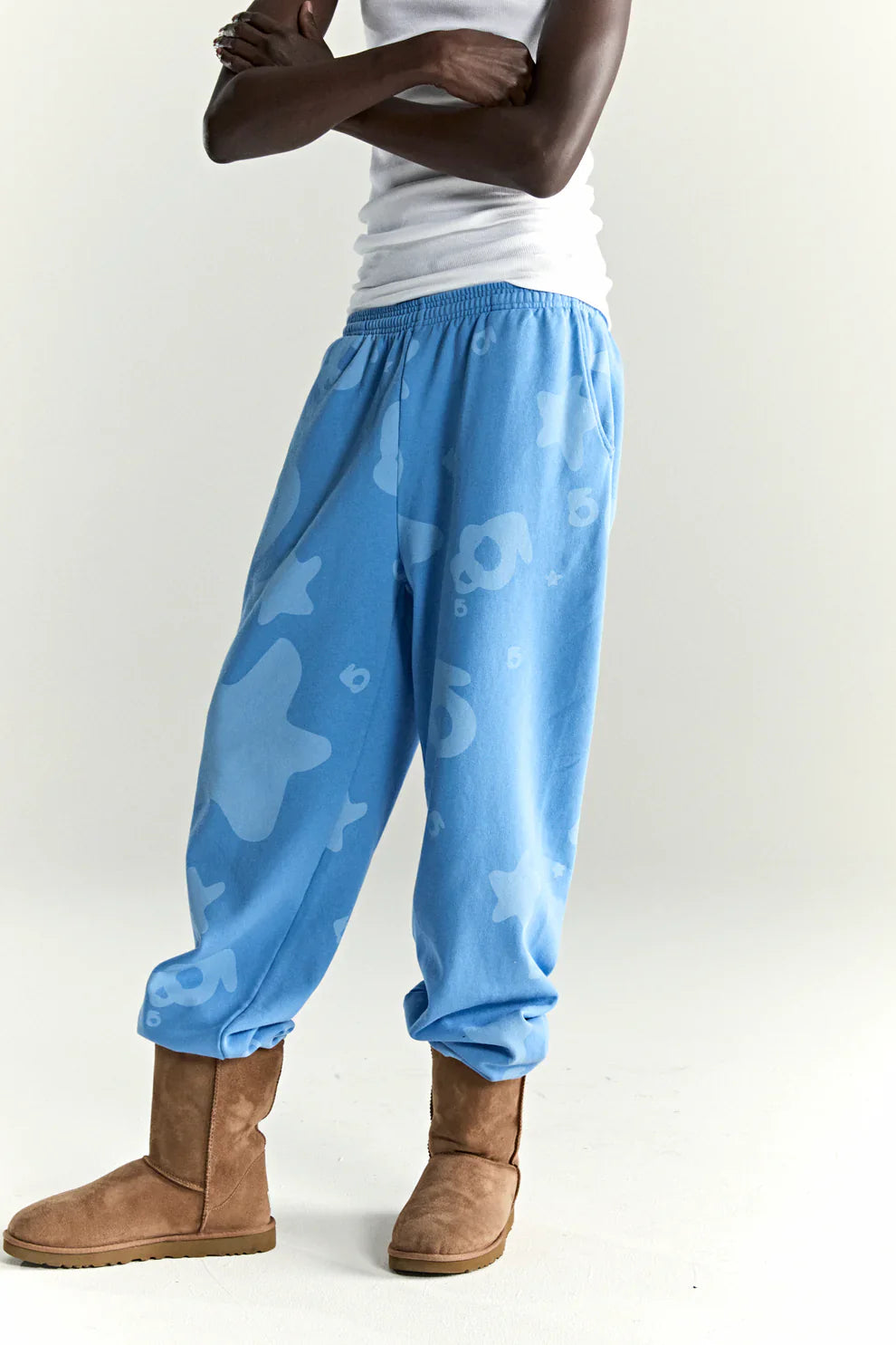 A person in a white tank top, Sp5der Light Blue All-Over Print Sweatpants 100% Cotton Unisex Streetwear Joggers, and brown UGG-style boots stands with arms crossed against a plain background.