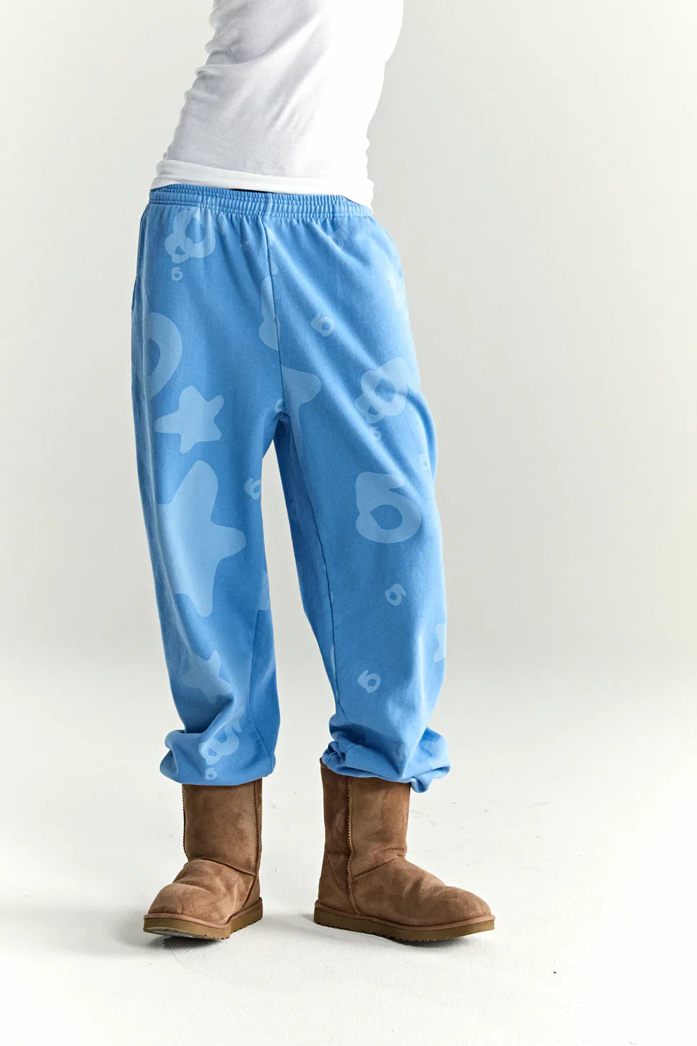 Wearing Sp5der Light Blue All-Over Print Sweatpants—100% cotton unisex streetwear joggers with abstract patterns—a person pairs them with brown suede boots and a white shirt, standing against a plain white background.