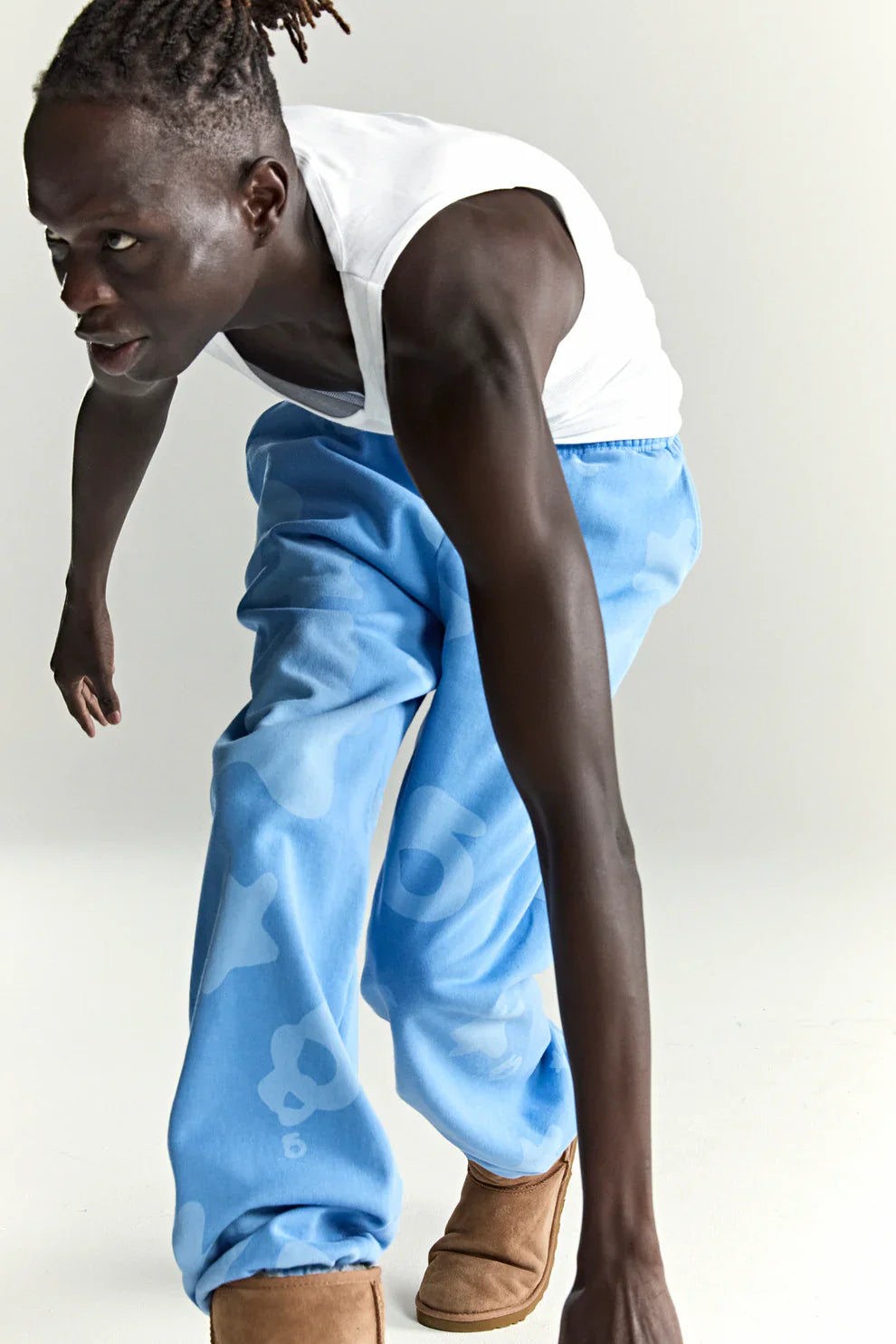 A person in a white tank top, Sp5der Light Blue All-Over Print 100% Cotton Unisex Streetwear Joggers, and tan boots is crouched forward in a dynamic pose against a plain white background.