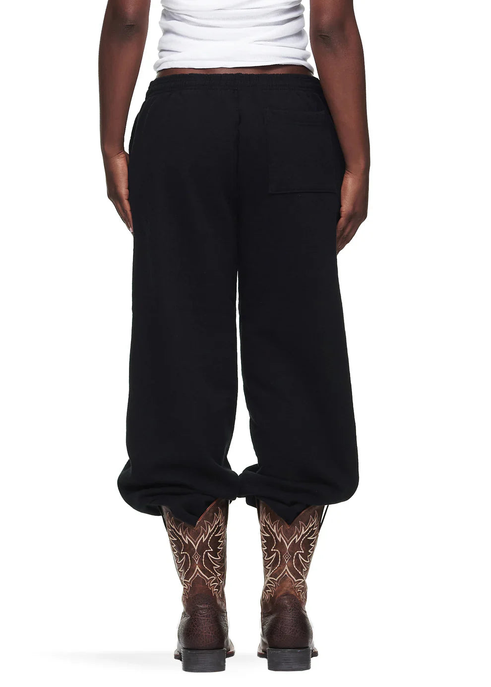 A person shown from the back is wearing Sp5der Sweatpants—unisex black cotton sweatpants with a front logo and star print—paired with a white tank top and brown cowboy boots featuring embroidered patterns.