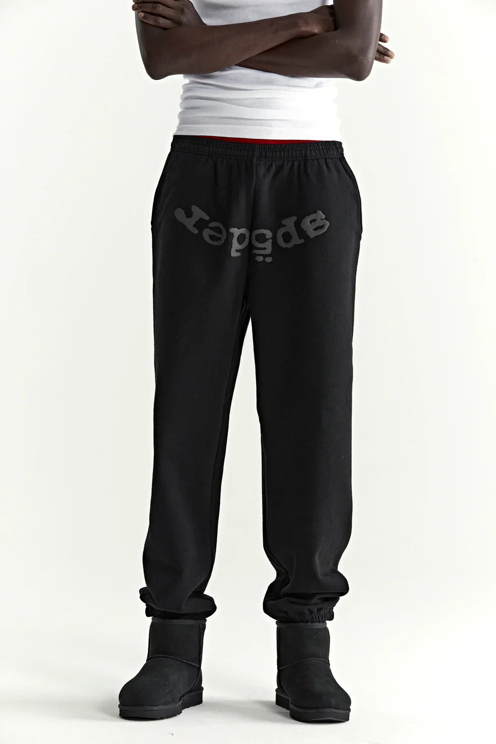 A person in a white tank top, Sp5der Black Tonal Logo Sweatpants (100% cotton, unisex streetwear joggers), and black boots stands against a light plain background with arms crossed, capturing a modern streetwear vibe.