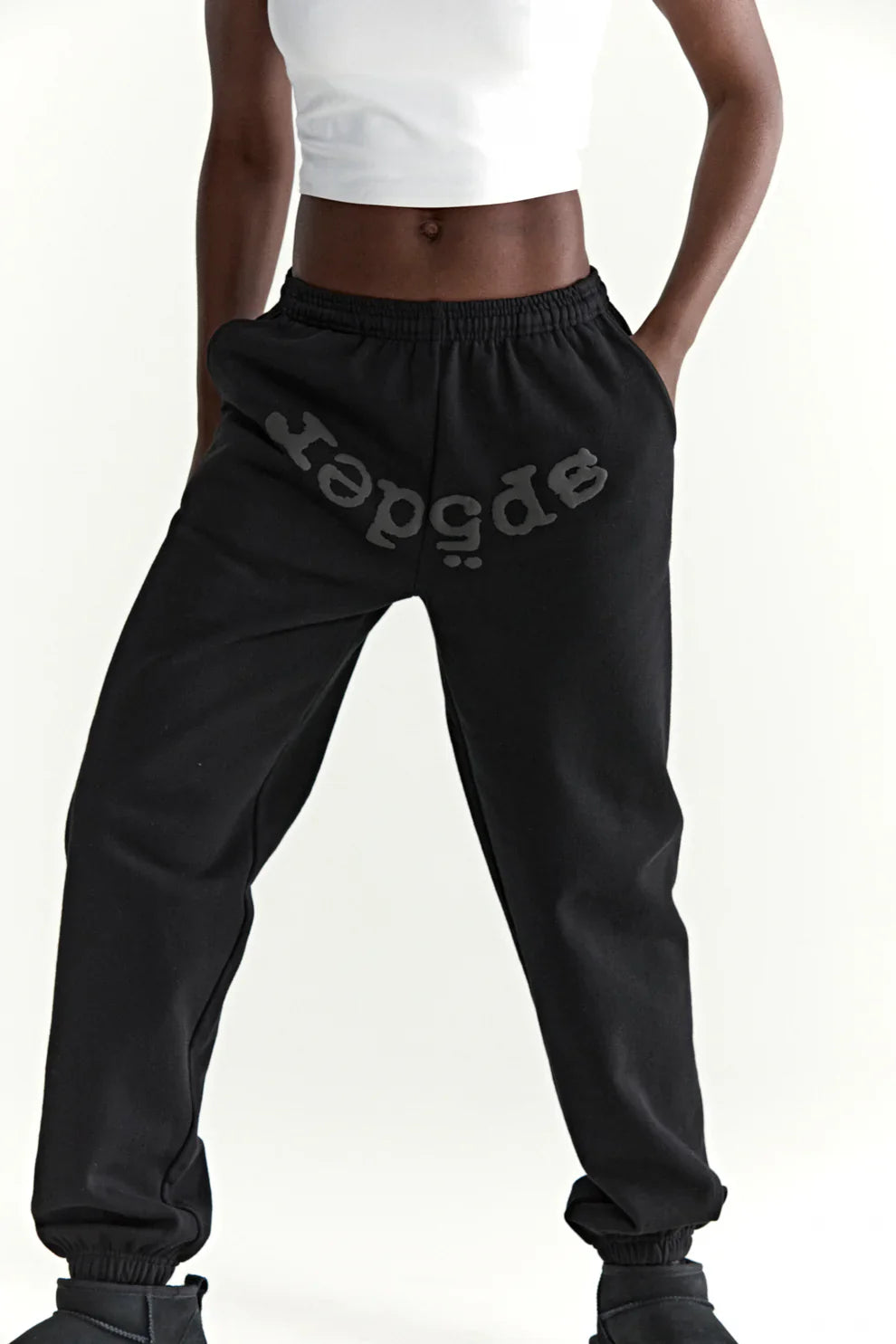 A person stands with hands in pockets against a plain white background, wearing a white crop top and Sp5der Black Tonal Logo Sweatpants—100% cotton unisex streetwear joggers—showcasing an effortless street style look.