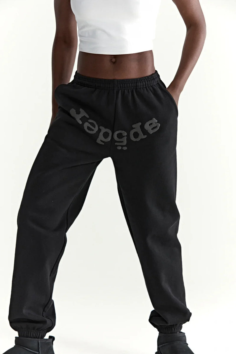 A person stands with hands in pockets against a plain white background, wearing a white crop top and Sp5der Black Tonal Logo Sweatpants—100% cotton unisex streetwear joggers—showcasing an effortless street style look.