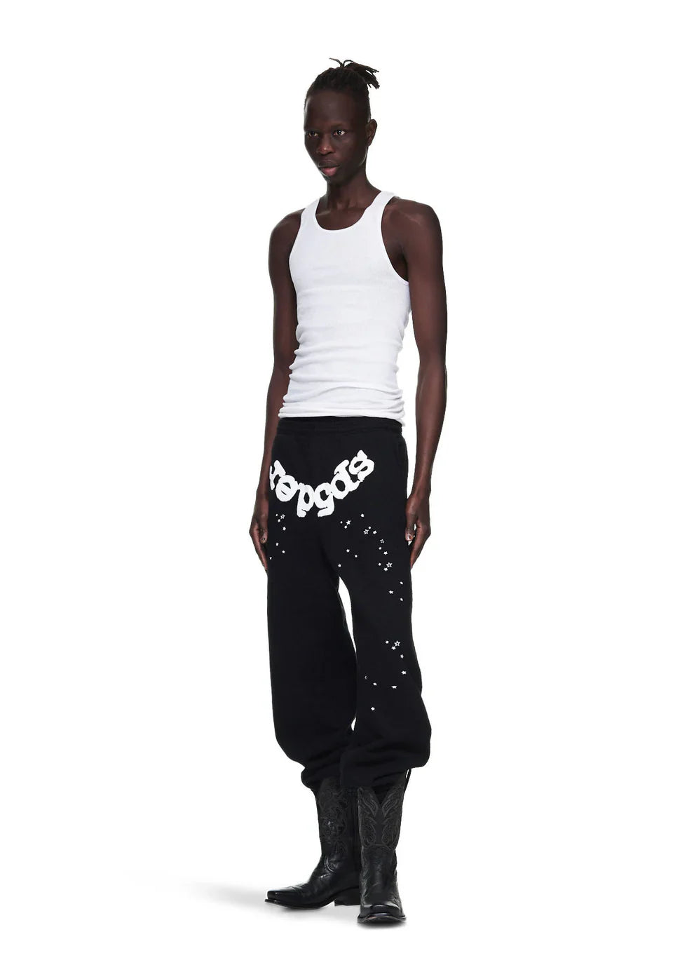 A person in a white tank top, Sp5der Sweatpants – Unisex Black Cotton Sweatpants with Front Logo and Star Print, and black cowboy boots stands against a plain white background.
