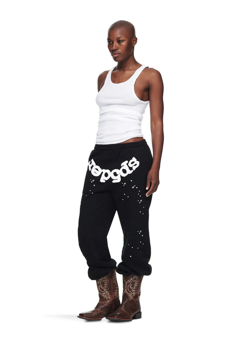A person with a shaved head wears a white tank top, Sp5der Unisex Black Cotton Sweatpants with front logo and star print, and brown cowboy boots, standing against a white background.