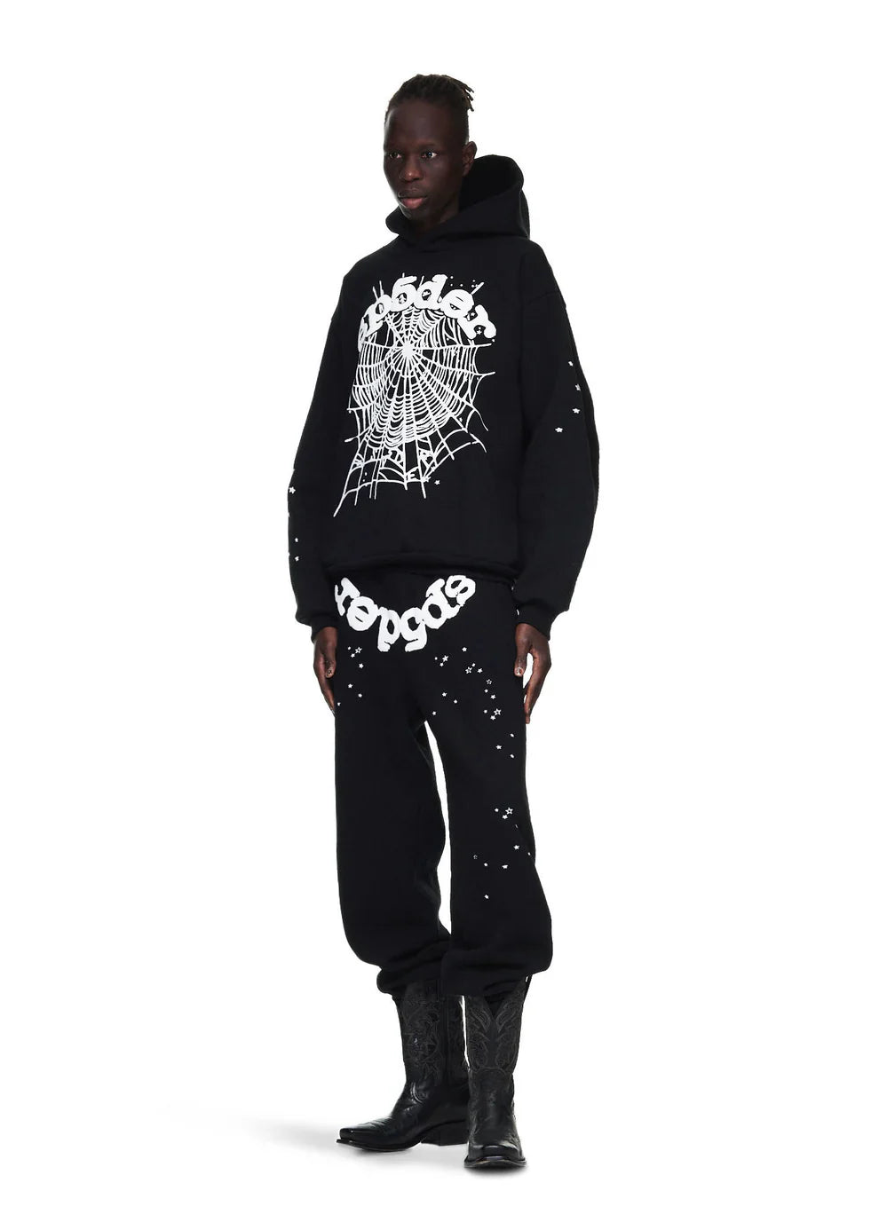 A person in a black hoodie with a white spiderweb design and Sp5der Sweatpants – Unisex Black Cotton Sweatpants with Front Logo and Star Print, paired with black cowboy boots, stands against a plain white background.