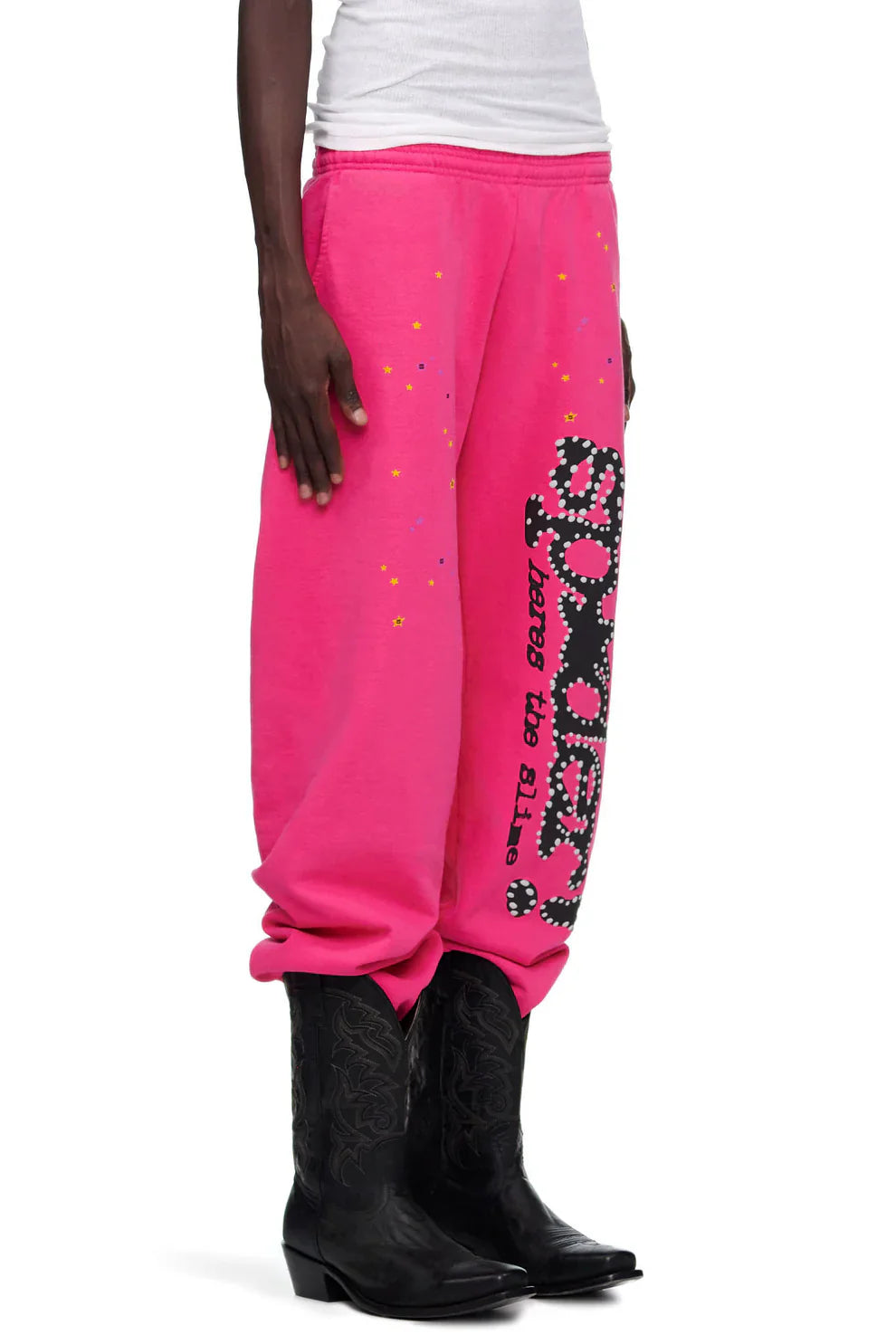 Someone wearing Sp5der Sweatpants—unisex pink cotton wide leg sweatpants with a graphic logo print—styled with black cowboy boots and a white top.