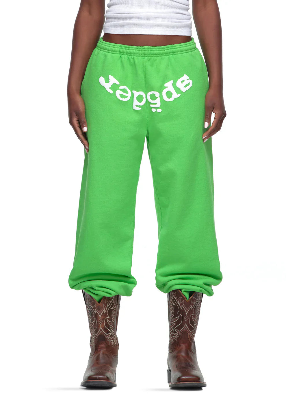 Someone wears Sp5der Sweatpants – Unisex Green Cotton Sweatpants with a front logo print, styled with a white tank top and brown embroidered cowboy boots. The sweatpants have gathered ankles for a relaxed streetwear vibe.