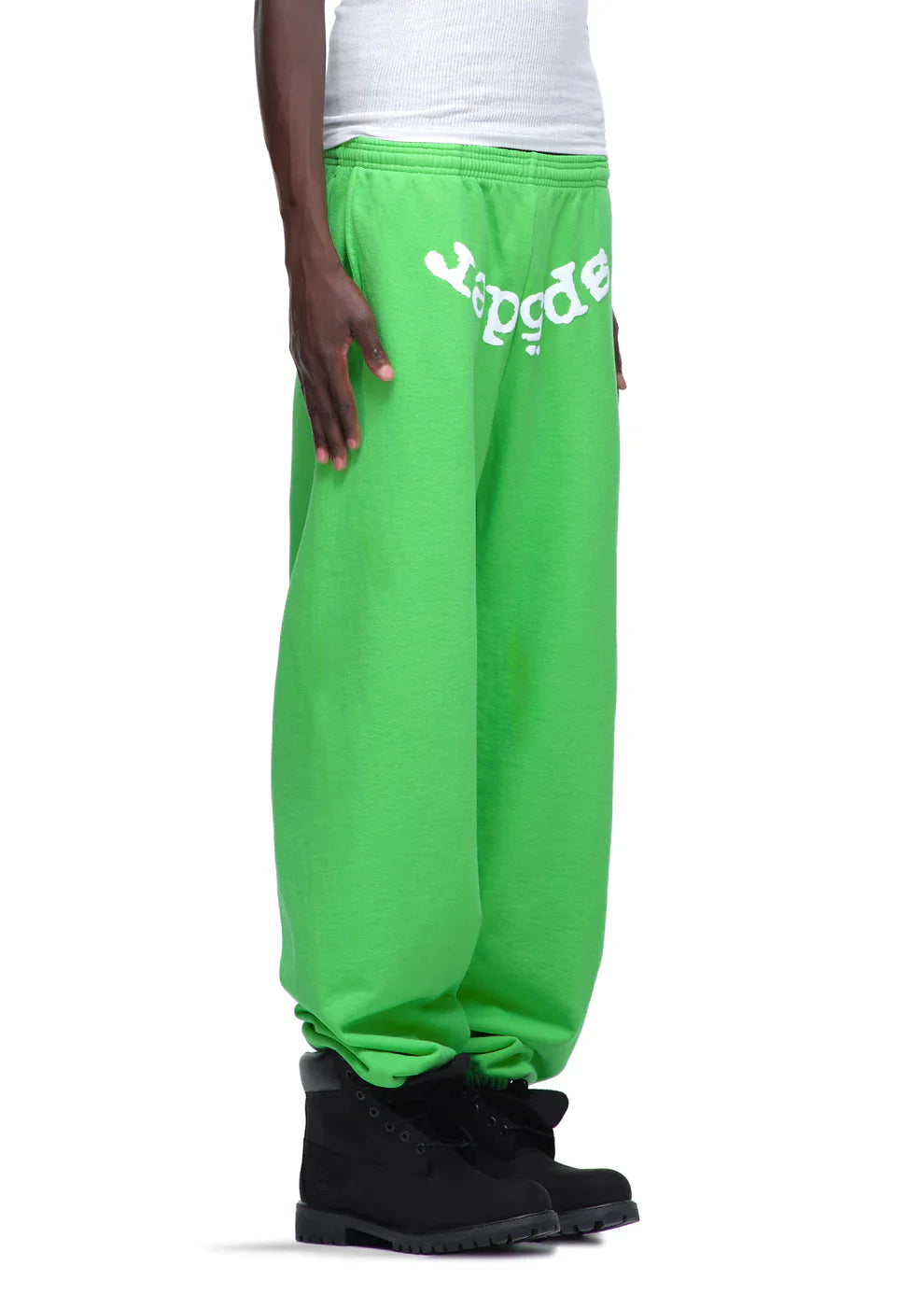 A person wearing Sp5der Sweatpants – Unisex Green Cotton Sweatpants with front logo print, paired with a white shirt and black boots, stands against a plain white background.