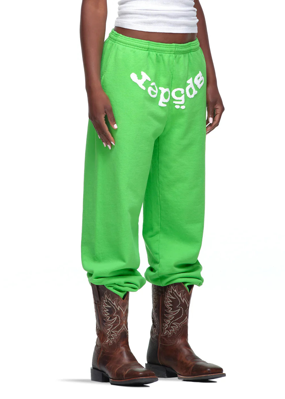 Someone wears Sp5der Sweatpants – Unisex Green Cotton Sweatpants with a front logo print, paired with a white tank top and brown cowboy boots. The joggers are rolled up above the boots as they pose against a white background.