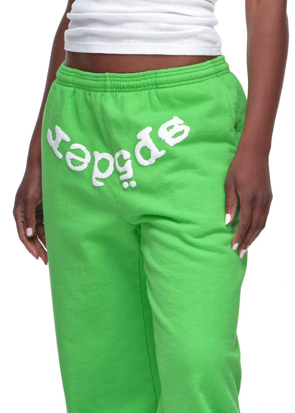 A person wears Sp5der Sweatpants – Unisex Green Cotton Sweatpants with a front logo print, paired with a white ribbed tank top. Only the torso and upper legs of this green streetwear look are visible.