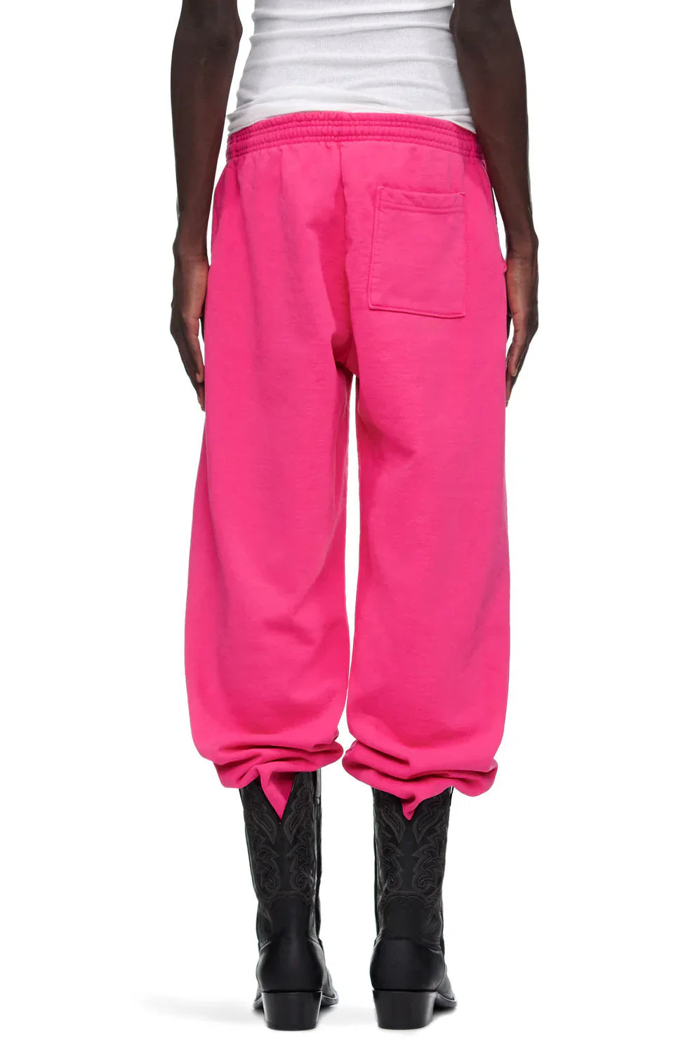 Someone stands with their back to the camera in Sp5der Sweatpants—unisex pink cotton wide leg sweatpants with a graphic logo print—styled with a white top and black cowboy boots for an effortless streetwear look.