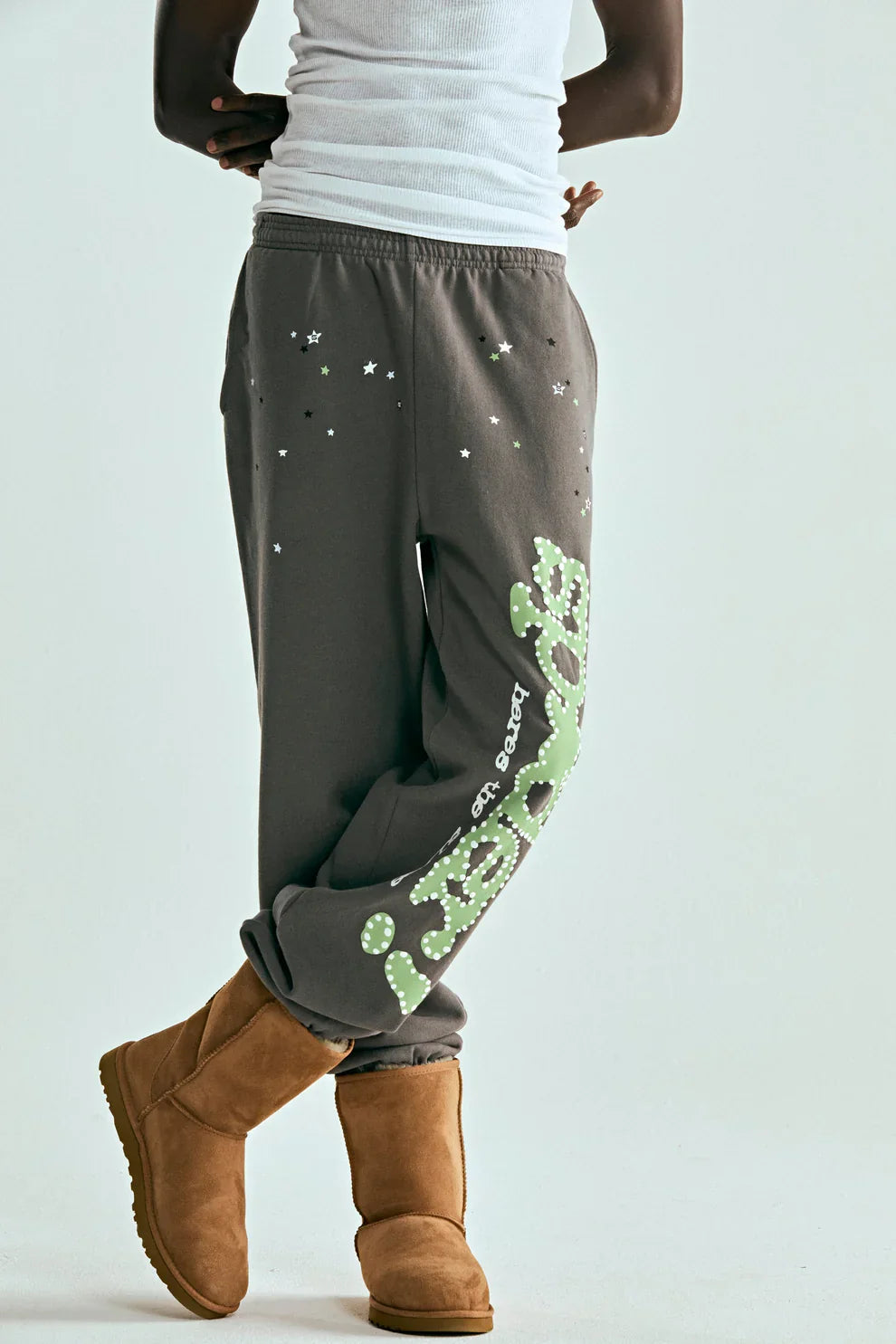 A person wearing a white tank top, Sp5der Sweatpants – Unisex Dark Grey Cotton Sweatpants with Green Side Logo and Star Print, and tan UGG-style boots stands with legs crossed against a plain light background.