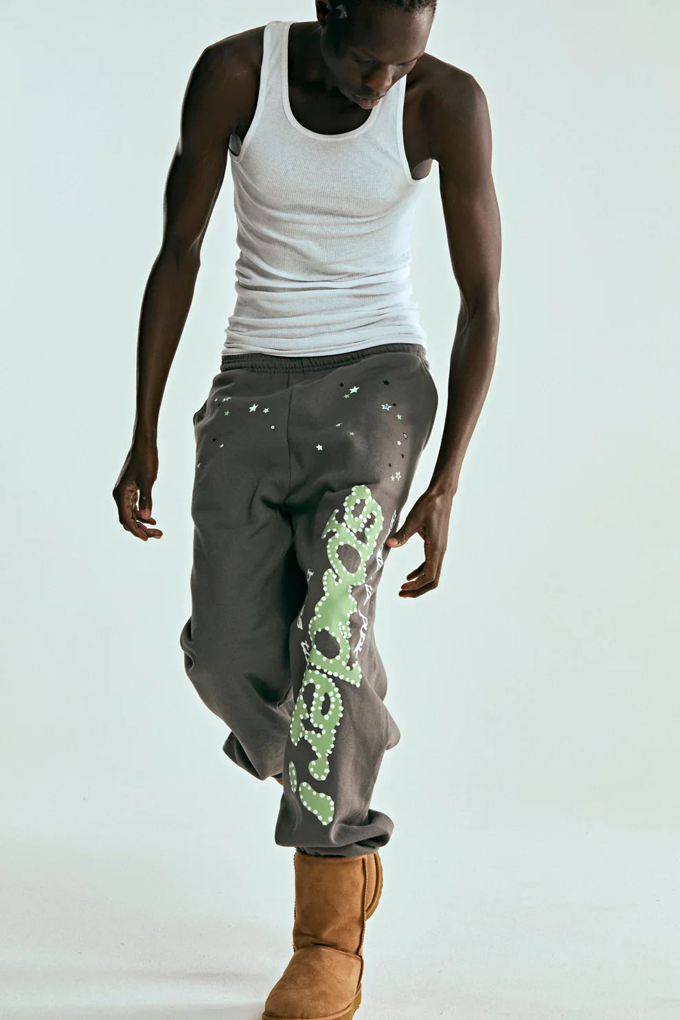 Dressed in a white tank top and Sp5der Sweatpants – unisex dark grey cotton sweatpants with a green side logo and star print – tan boots, the person stands against a plain light background, looking down with one hand at their side.
