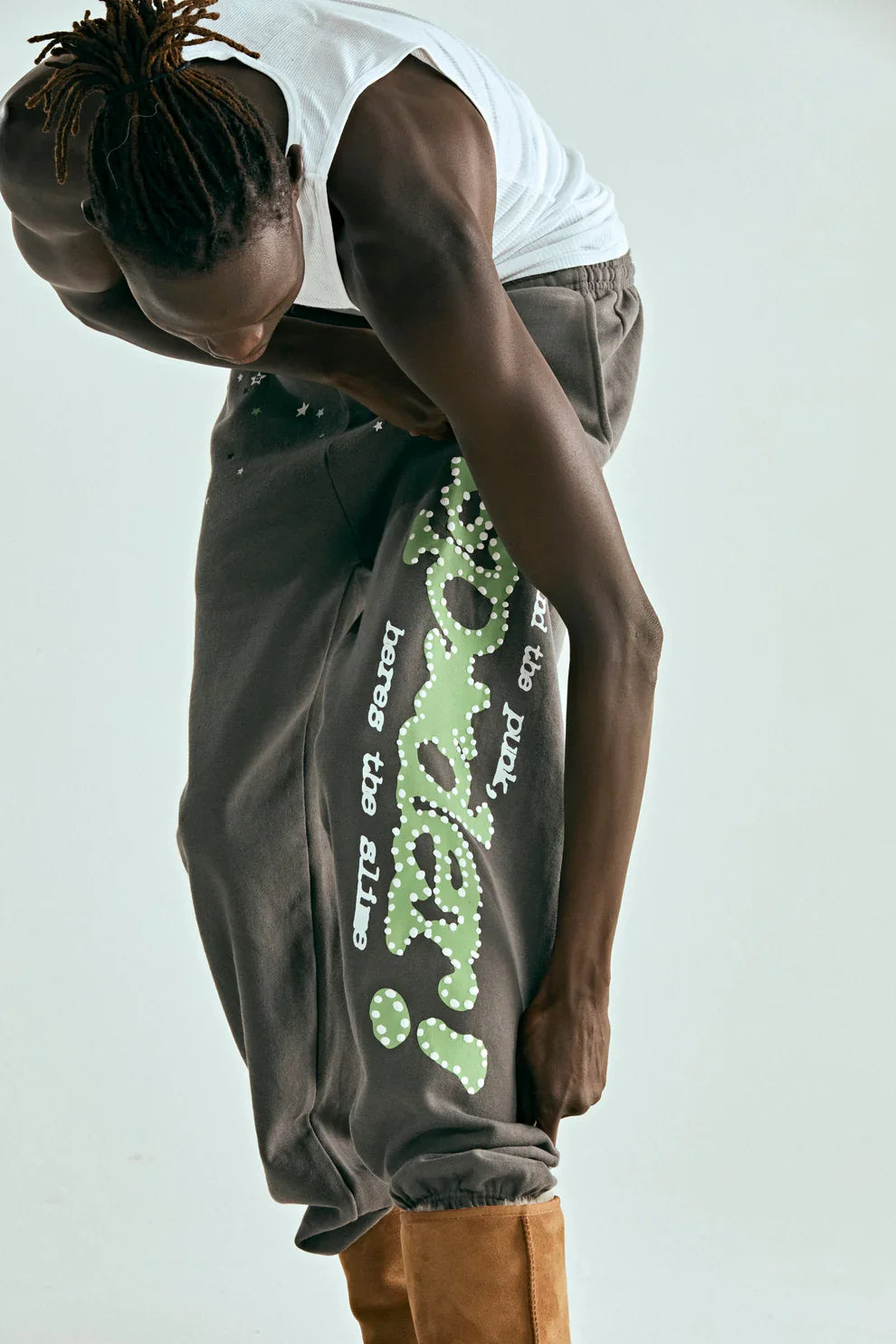 A person in a white tank top and brown pants bends forward, holding their pant leg to showcase Sp5der Sweatpants—unisex dark grey cotton sweatpants featuring a green side logo and star print—against a simple light background. They have twisted hair.
