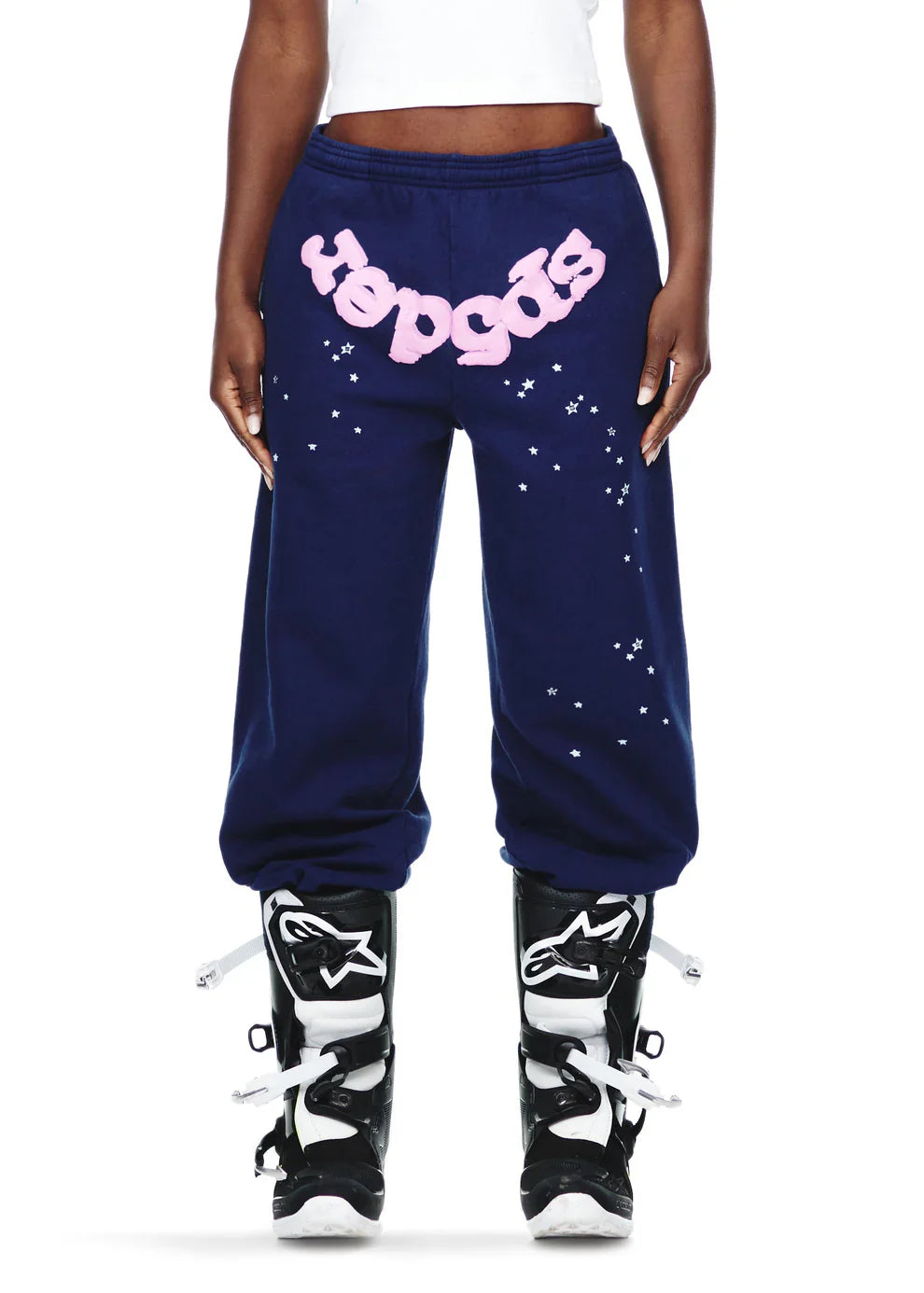 A person wears Sp5der Sweatpants—unisex navy blue cotton with a pink front logo and star print—paired with black-and-white motocross boots. The sweatpants sit at the waist, revealing a visible midriff.
