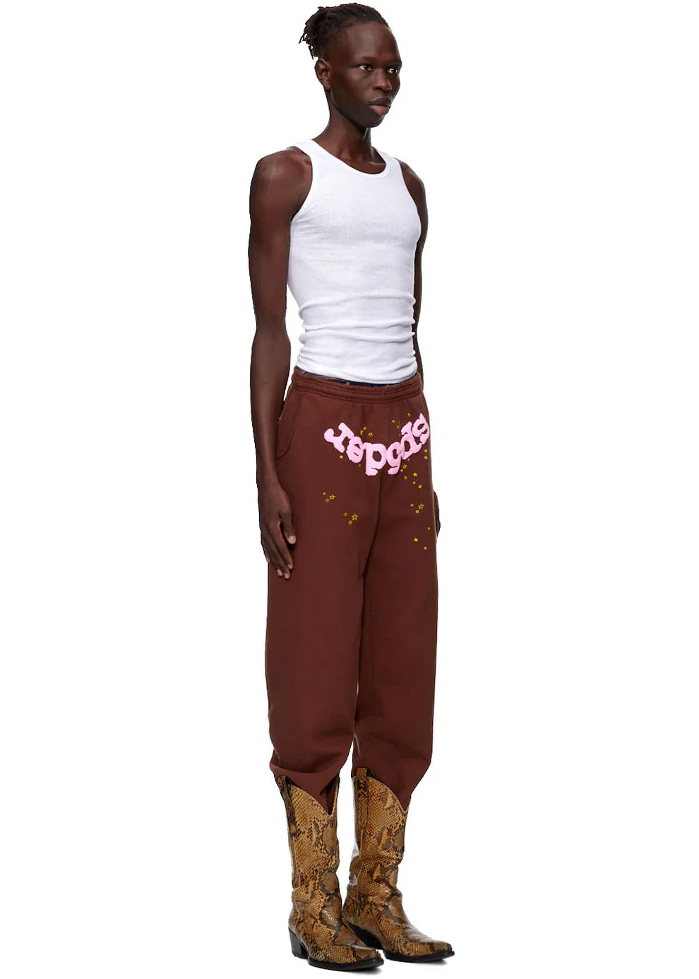 A person stands sideways in a white tank top, tan snakeskin cowboy boots, and Sp5der Sweatpants—unisex brown cotton sweatpants with a pink front logo and star print—against a white background for streetwear style.