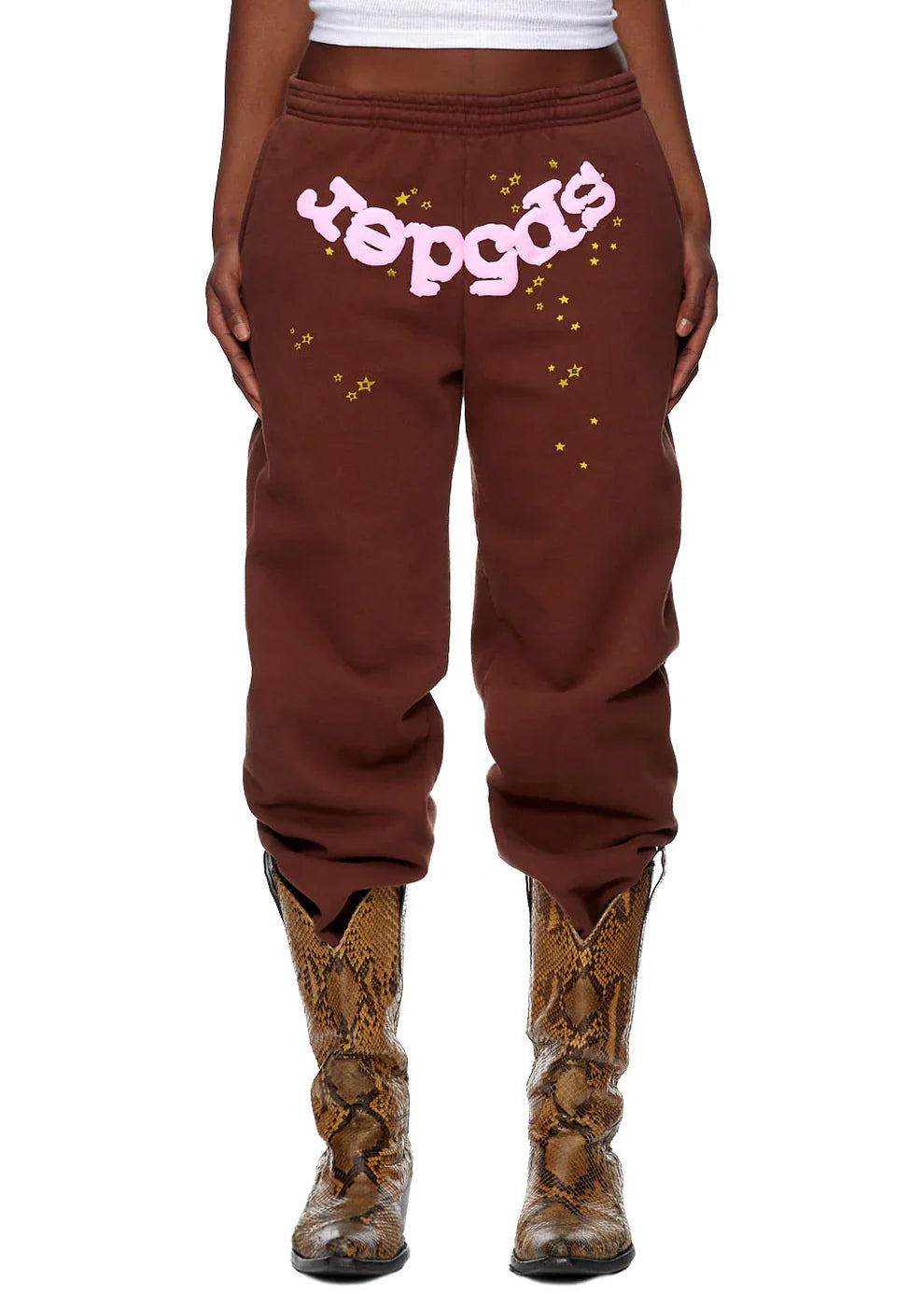 A person wears Sp5der Sweatpants—unisex brown cotton sweatpants with a pink front logo and star print—paired with brown snakeskin cowboy boots. The cropped image highlights a trendy streetwear look focused on the lower torso and legs.