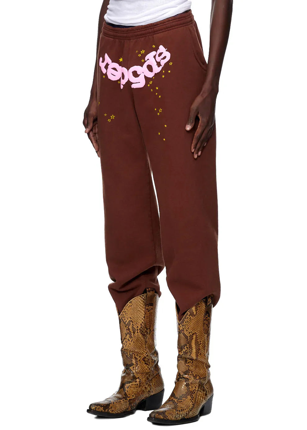 A person wears Sp5der unisex brown cotton sweatpants featuring a pink front logo and star print, styled with a white top and brown snakeskin cowboy boots. Hands are tucked into the pockets for a cool streetwear look.