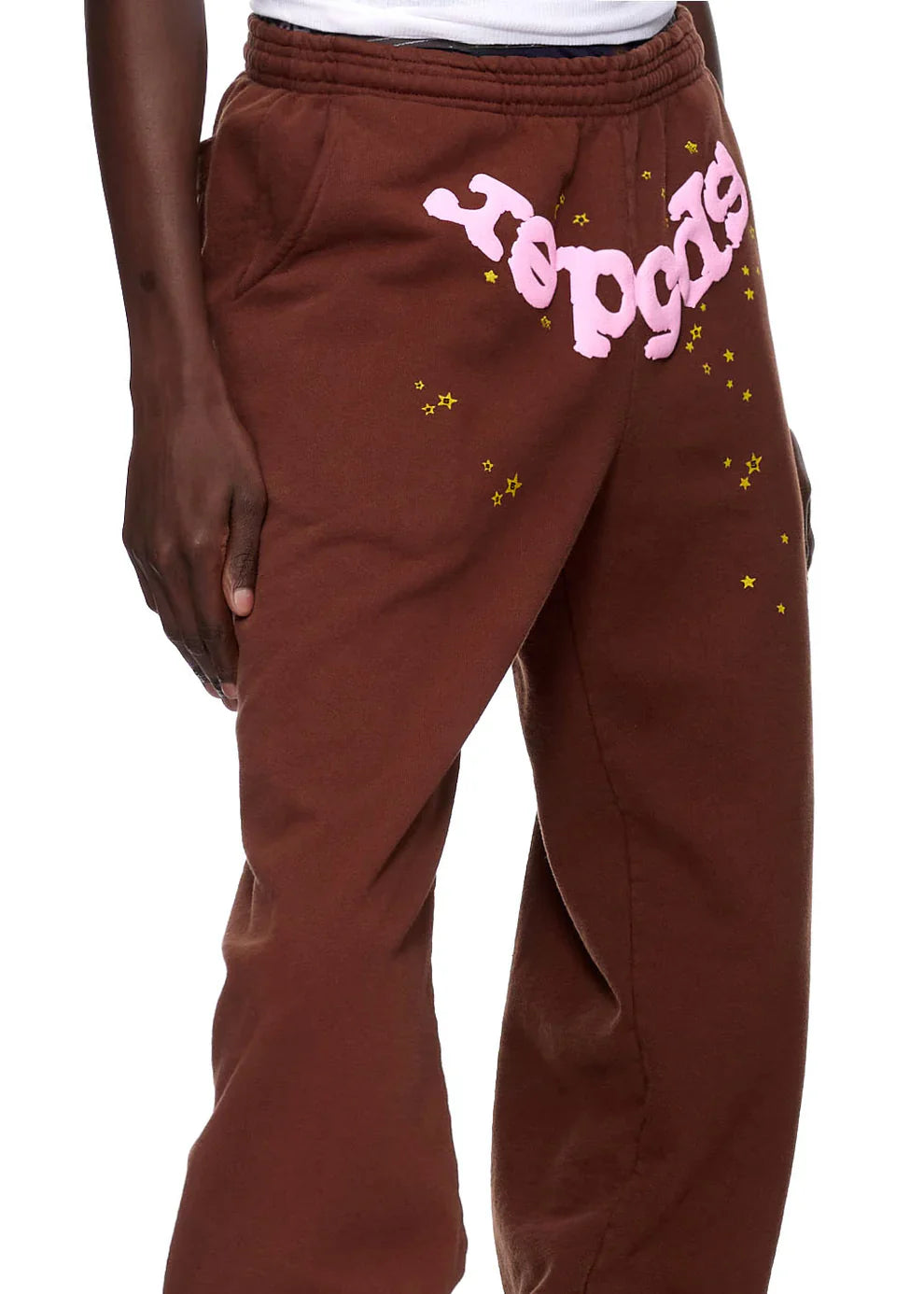 A person in Sp5der Sweatpants—unisex brown cotton sweatpants with a pink front logo and star print—creates an eye-catching streetwear look, styling these joggers with one hand by their side.