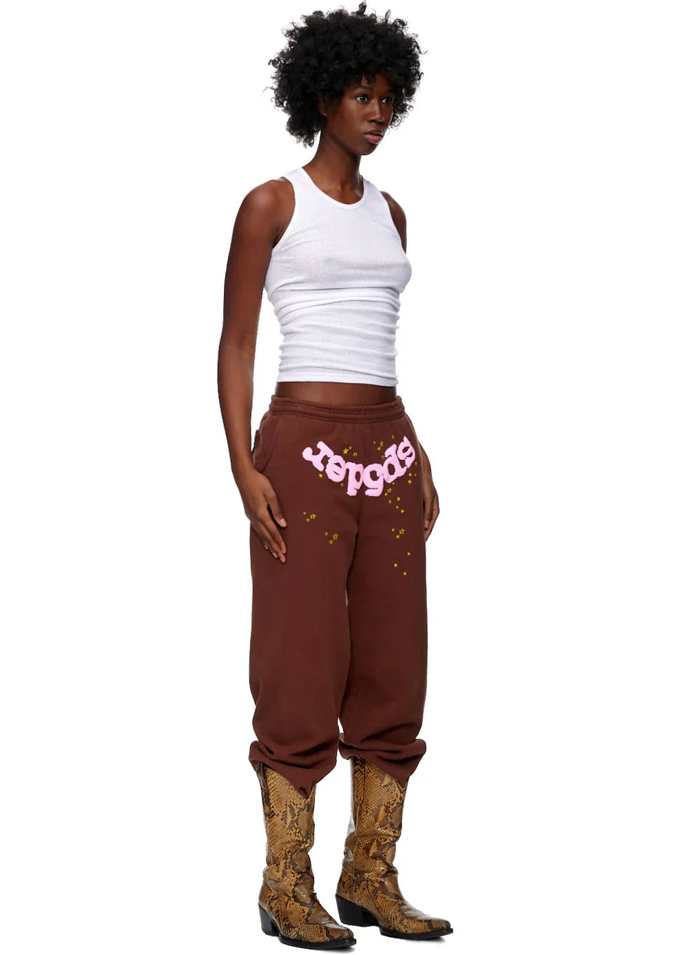 A person with short curly hair wears a white tank top, Sp5der Sweatpants—unisex brown cotton sweatpants with a pink front logo and star print—and tan snakeskin cowboy boots against a plain white background.