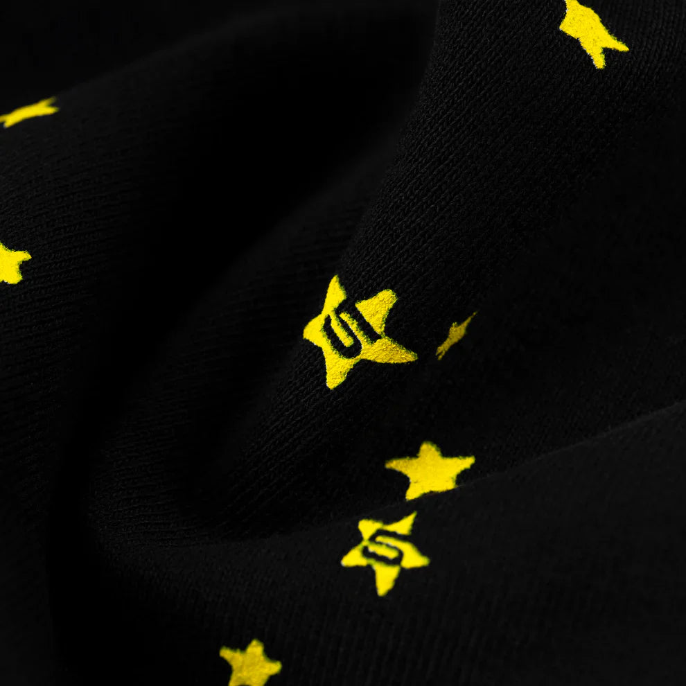 Close-up of Sp5der Sweatpants—Unisex Black Cotton Sweatpants with a flame logo on the side and yellow star print, some with a black "S." Slightly folded fabric adds soft shadows and texture, ideal for trendy streetwear joggers.