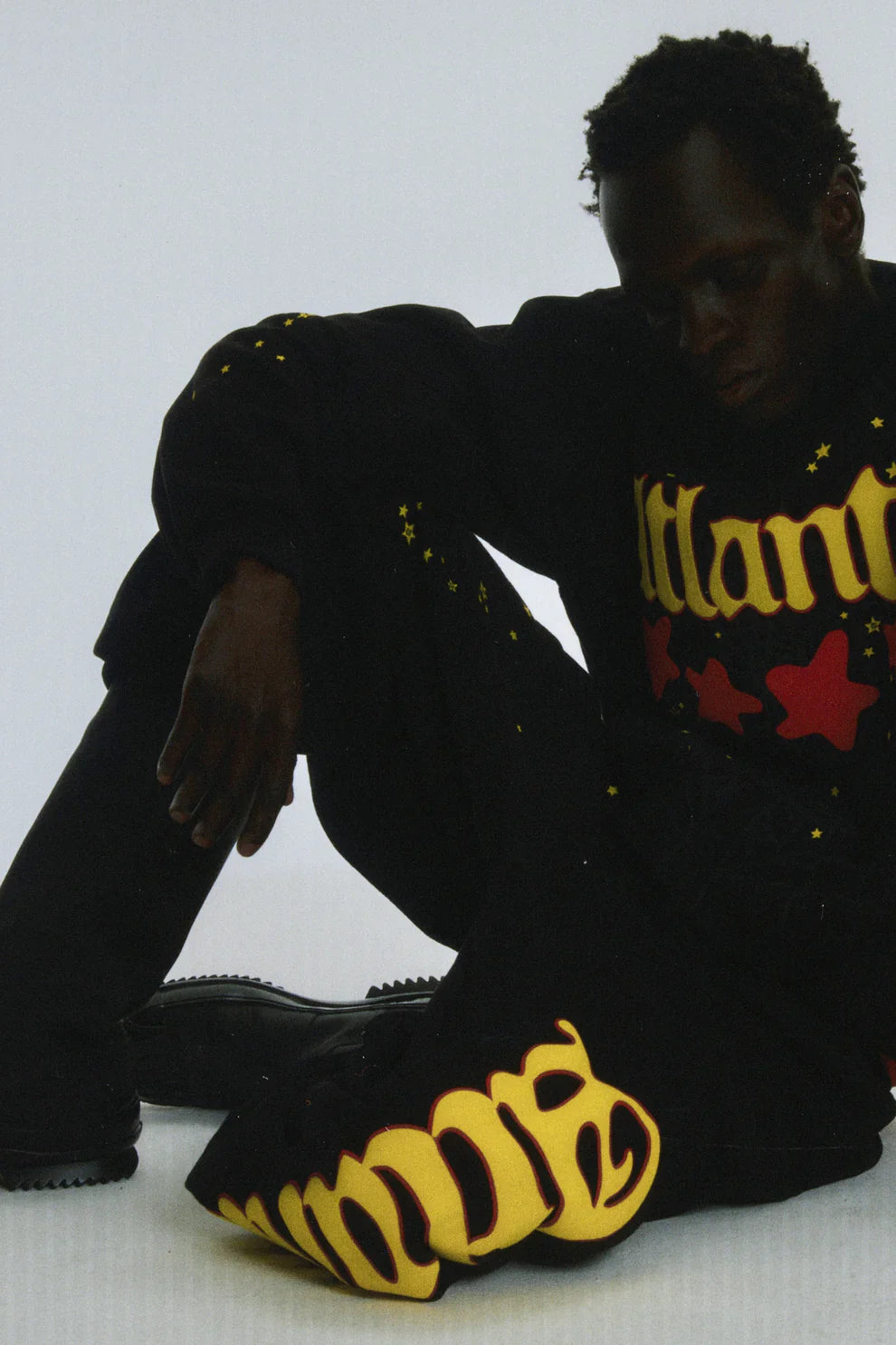 Against a light background, a person bows their head, face mostly shadowed, wearing unisex Sp5der Sweatpants—black cotton with flame side logo and star print. Bold yellow and red graphic text decorates the streetwear joggers.