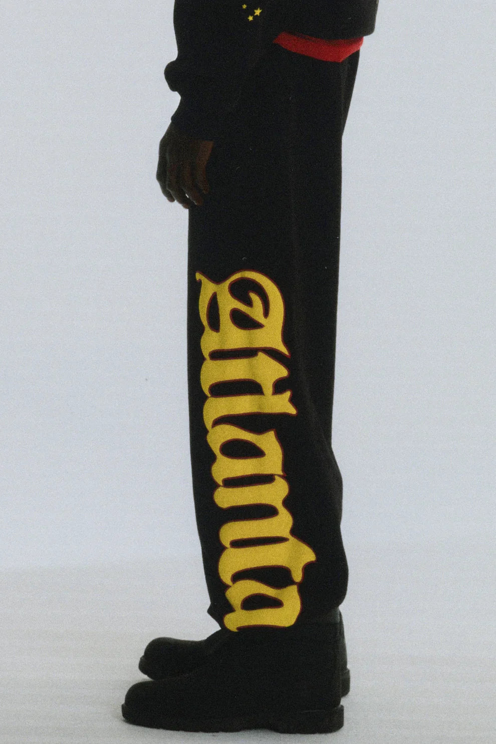 A person wears Sp5der Sweatpants – Unisex Black Cotton Sweatpants with a flame side logo and star print, paired with black shoes. The photo shows only the lower body against a plain background, highlighting streetwear joggers style.