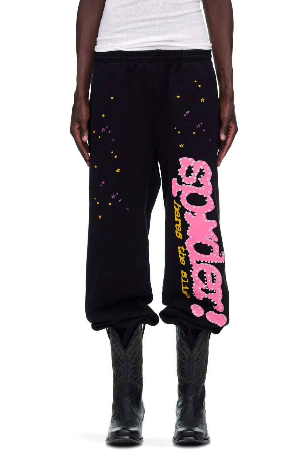 A person is shown from the waist down wearing Sp5der Sweatpants—unisex black cotton joggers with a pink graphic logo and star print—paired with a white tank top and black cowboy boots.