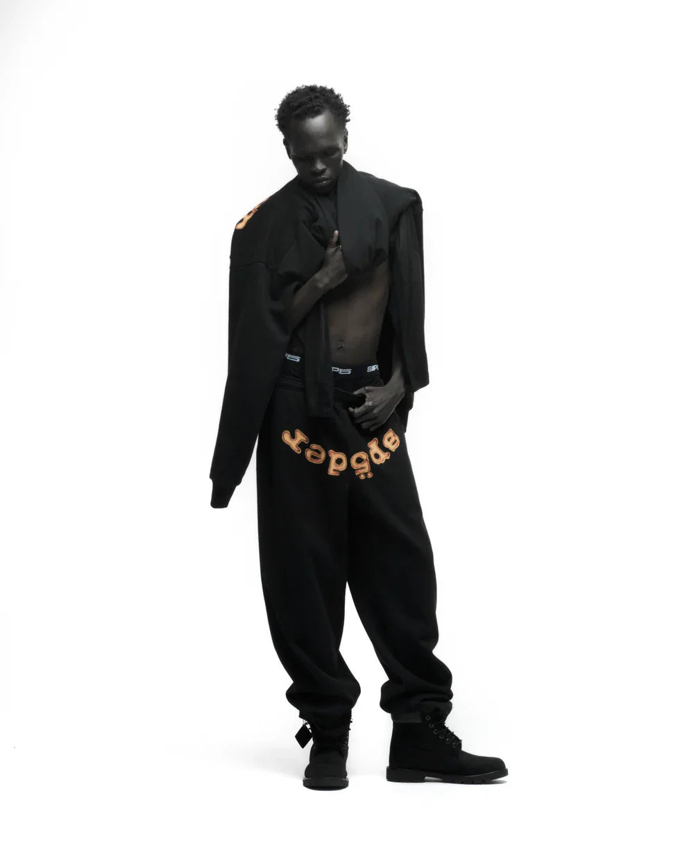 A person poses against a white background, wearing Sp5der Sweatpants – unisex black cotton sweatpants with a flame front logo print, paired with black boots and a partially draped black jacket exposing their chest.