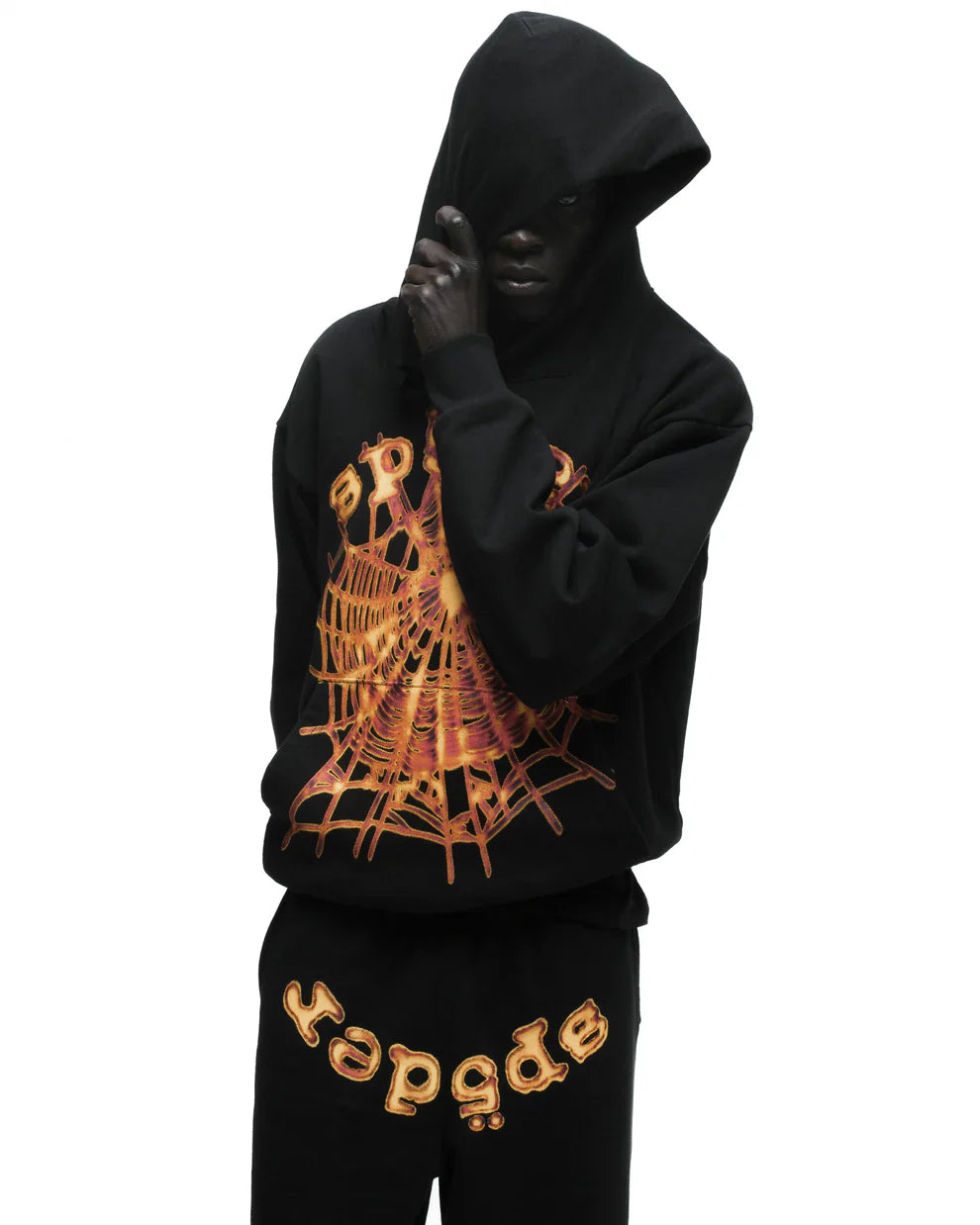 A person in a black hoodie with an orange spiderweb design and Sp5der Sweatpants—unisex black cotton sweatpants with a flame front logo print—stands against a white background, partially hiding their face with the hood for standout streetwear style.