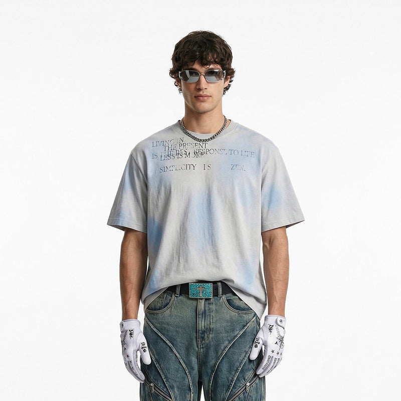 Vintage spray-dyed oversized tee with faded blue-gray print, styled on a model with jeans, gloves, and sunglasses