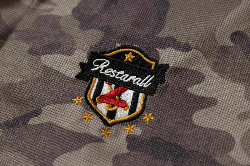 Close-up of embroidered restaurant crest on purple camo fabric, featuring a shield logo with stars on a short-sleeve tee