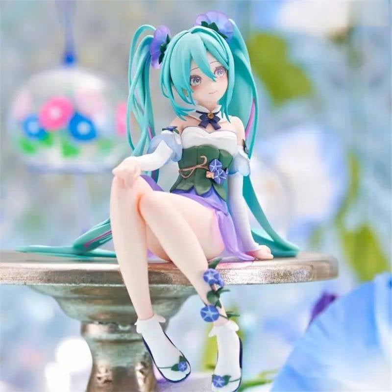 A Hatsune Miku Fairy 13.5cm PVC Figure in a sitting pose is displayed on a round table. The anime character has long blue-green hair and wears a pastel purple and green outfit with flower motifs, against a blurred background of soft blues and greens.