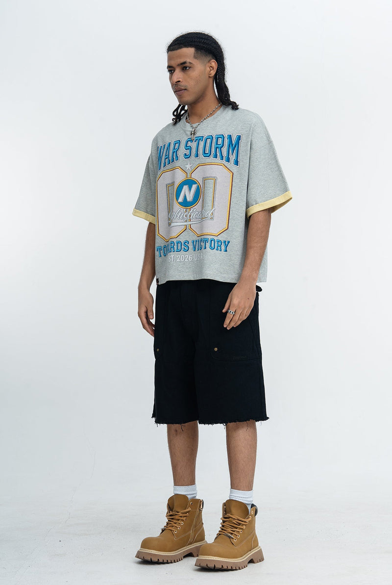 Model wearing a gray War Storm varsity graphic tee with blue lettering, black shorts, and tan lace-up boots on a white background