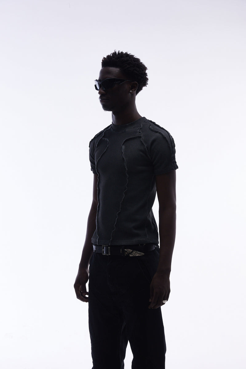 Model wearing a dark raw seam compression tee with deconstructed stitching, paired with black pants and sunglasses