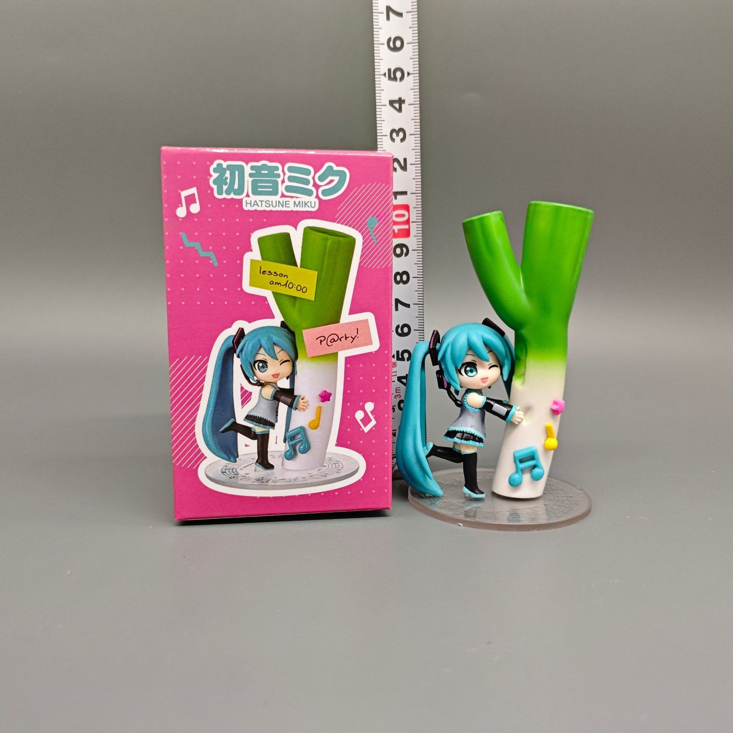 A Chibi Hatsune Miku figurine, featuring blue twin tails and holding a green onion, stands next to a matching pink box with Japanese text. A ruler is positioned behind the 12 cm PVC figure from the Hatsune Miku brand.