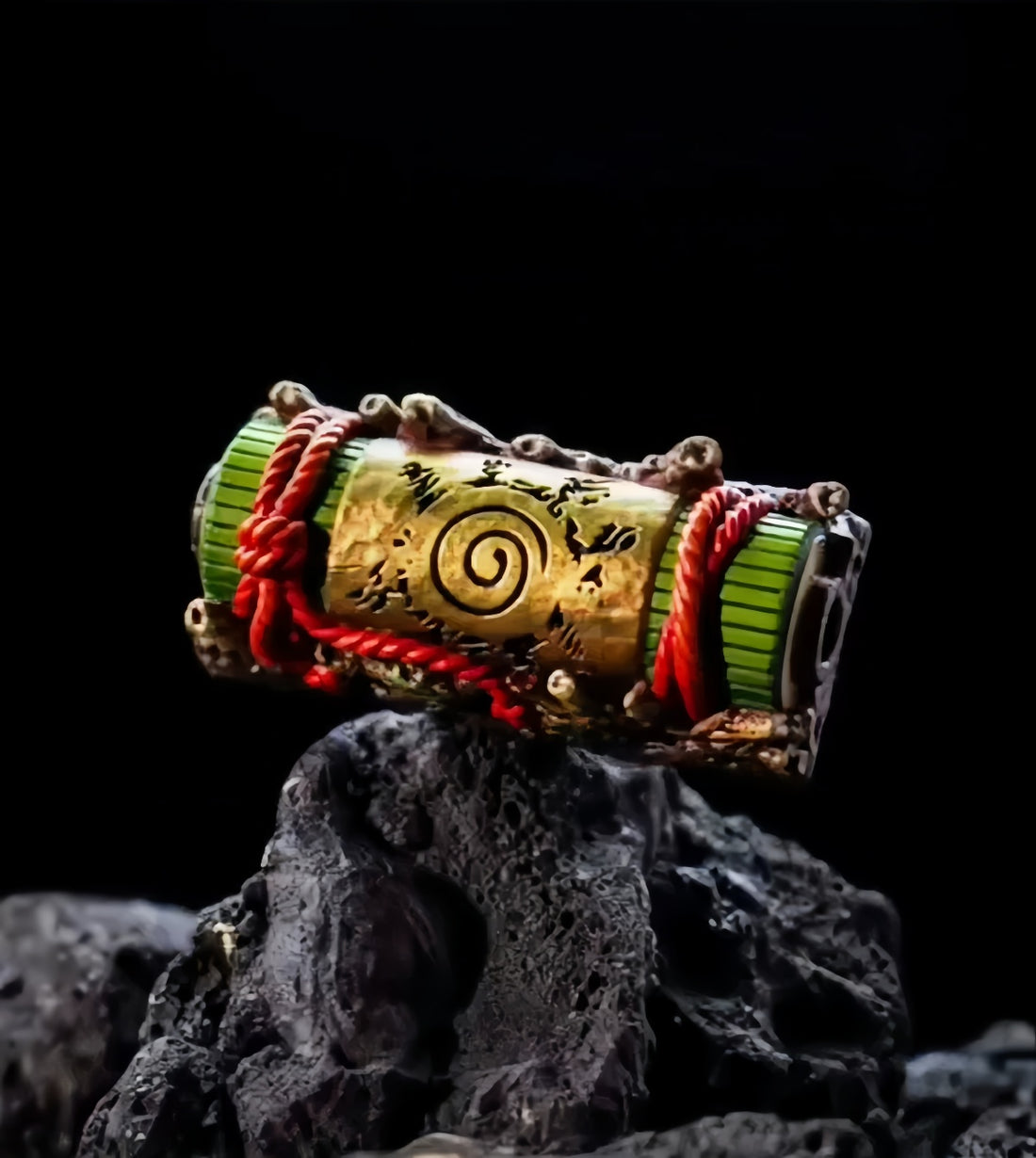 A decorative scroll featuring a green top and gold casing with a spiral design sits on dark terrain, resembling the intricate Ninja Keycaps Set by Naruto. Red string enhances its mystique, echoing the stealth of a shinobi.