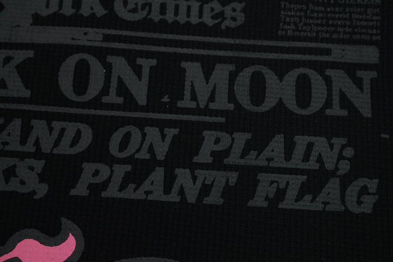 Close-up of a black waffle tee with faded vintage newspaper-style text and a small pink graphic accent