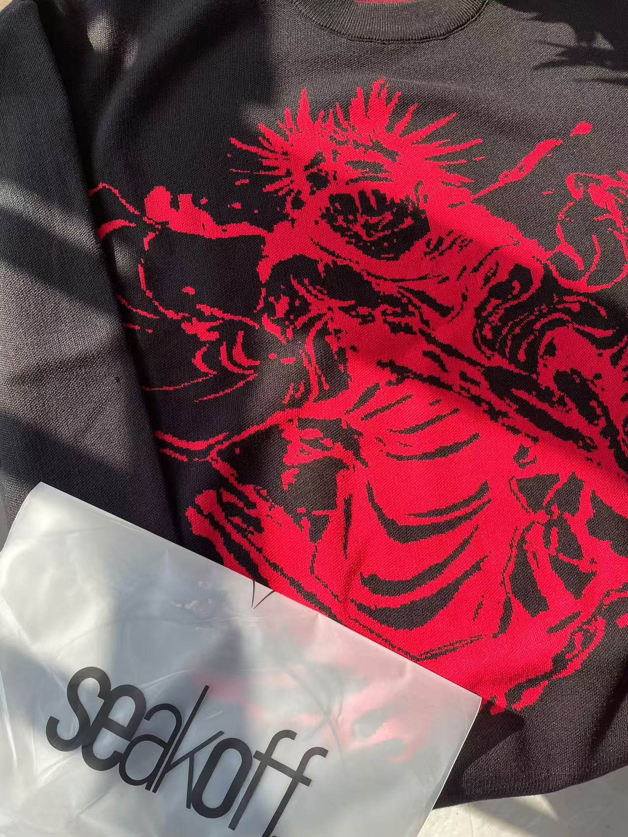 A Jujutsu Kaisen Inspired Black Sweatshirt features a red cursed energy design and hints of anime streetwear style. A transparent plastic bag below reads &quot;seekoff,&quot; partially shadowed to suggest hidden energy.