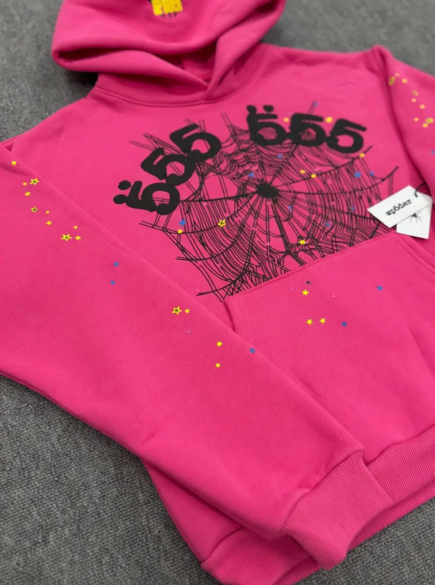 The Sp5der 555 Web Pink Hoodie Streetwear Pullover features a bold black spider web graphic and "Sp5der 555" on the chest, with yellow and blue stars and dots on the sleeves. It is shown laid flat on a gray surface.