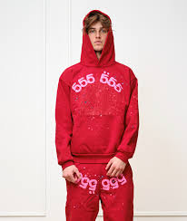 A person wears the Sp5der “555” Web Hoodie in red & pink, a 440GSM heavyweight oversized streetwear pullover, with matching pants featuring pink numbers, standing against a plain white background.