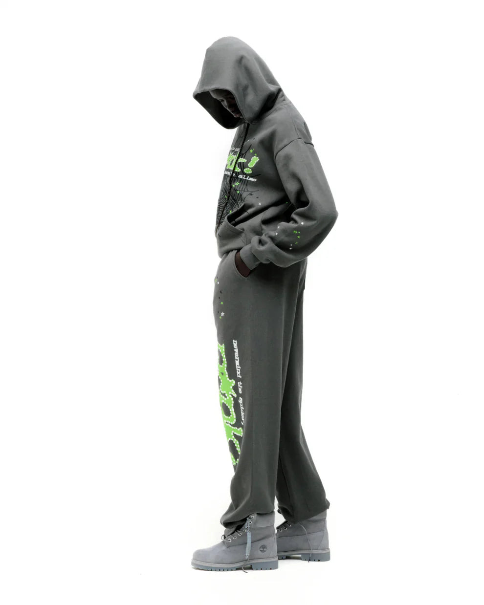 A person stands sideways with head down, hands in pockets, wearing the olive green Sp5der “P!nk! Hares the Slime” 440GSM heavyweight spider-web pullover hoodie and gray boots, striking a classic streetwear pose against a white background.