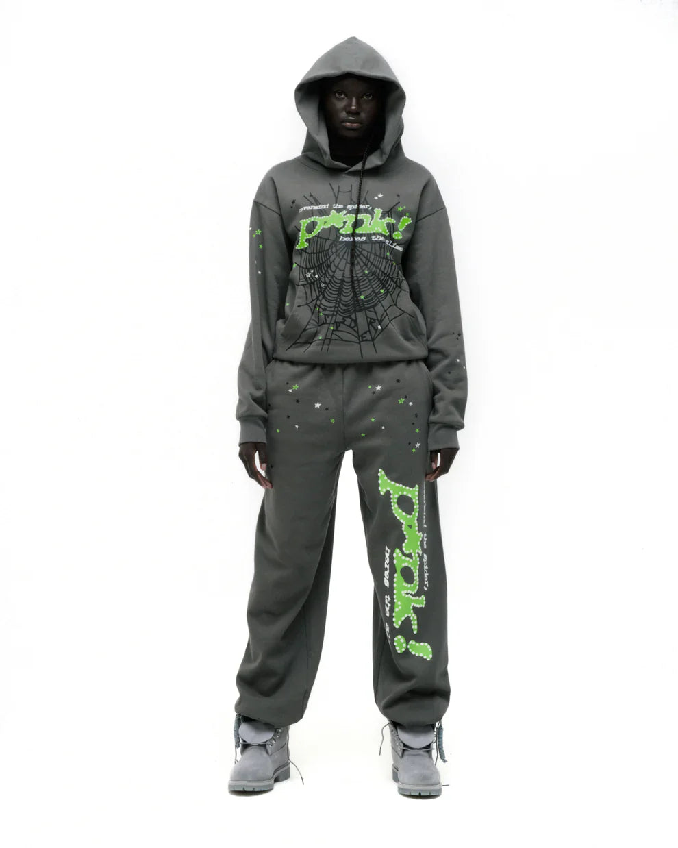 A person wears the Sp5der “P!nk! Hares the Slime” Olive Green 440GSM Heavyweight Spider-Web Pullover hoodie with matching sweatpants, both featuring neon green text and a spiderweb graphic, standing against a white background in bold streetwear style.