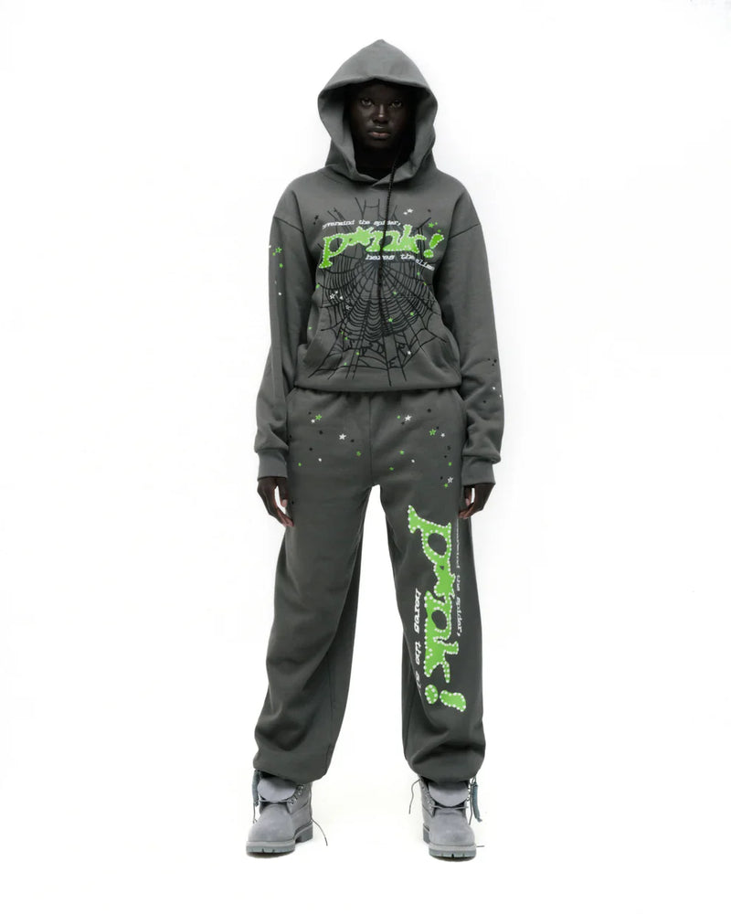 A person wears the Sp5der “P!nk! Hares the Slime” Olive Green 440GSM Heavyweight Spider-Web Pullover hoodie with matching sweatpants, both featuring neon green text and a spiderweb graphic, standing against a white background in bold streetwear style.