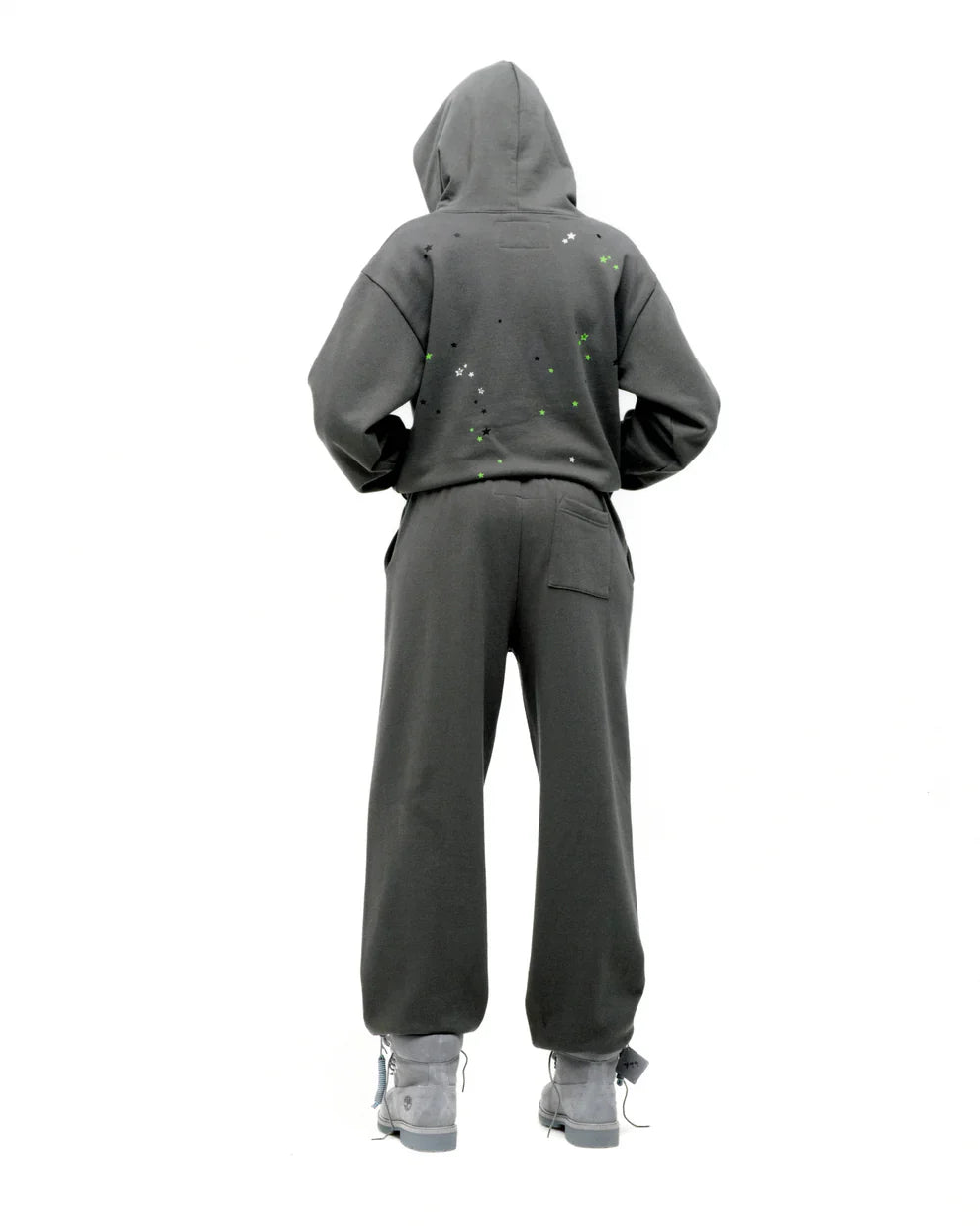 A person with their back to the camera wears an olive green Sp5der “P!nk! Hares the Slime” 440GSM heavyweight spider-web pullover hoodie, paired with matching pants and gray shoes, set against a plain white background.