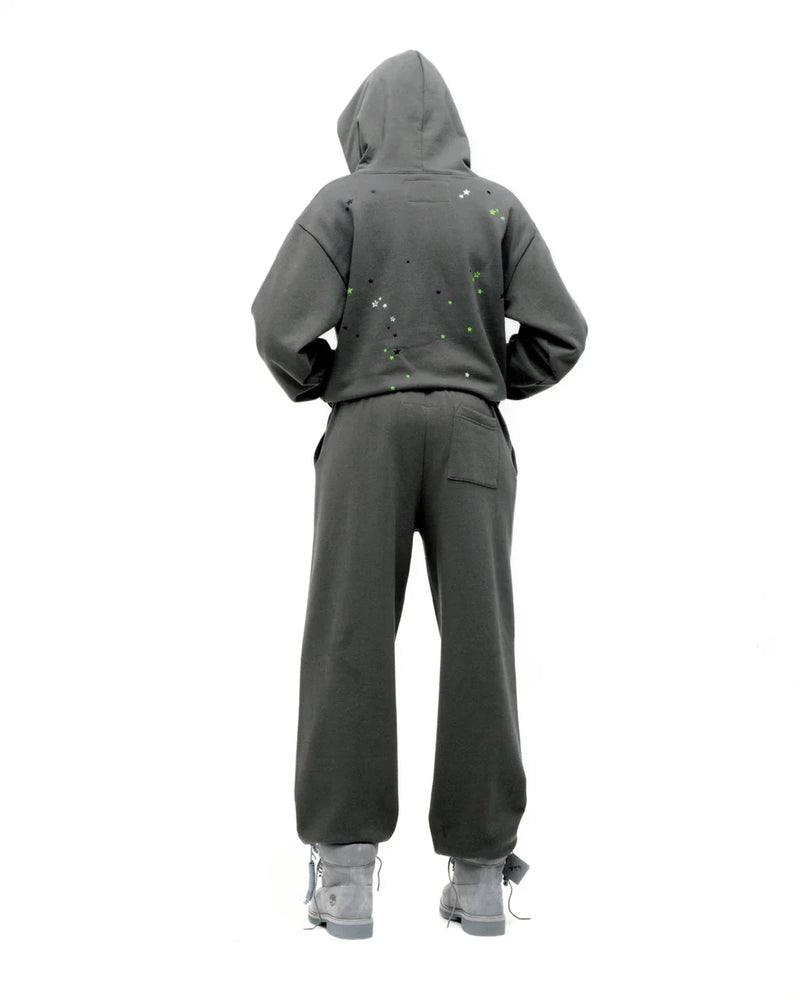A person with their back to the camera wears an olive green Sp5der “P!nk! Hares the Slime” 440GSM heavyweight spider-web pullover hoodie, paired with matching pants and gray shoes, set against a plain white background.